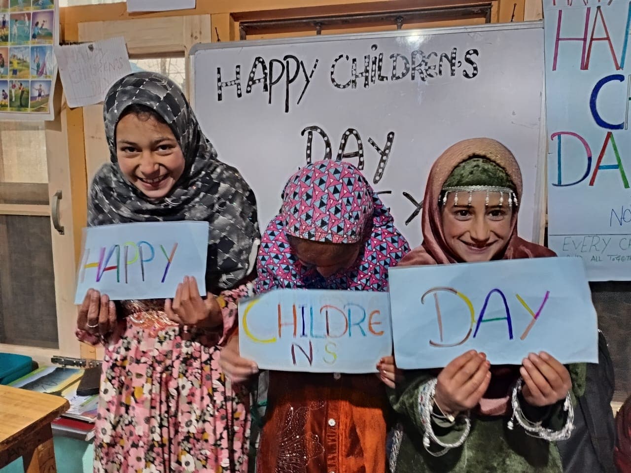 Three girls holding hand-coloured Happy Children's Day signs in a classroom