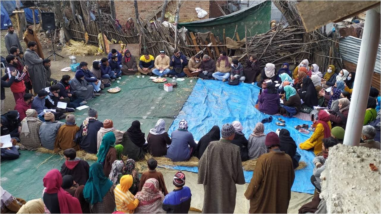 A large open-air community meeting with residents seated on mats in a village courtyard