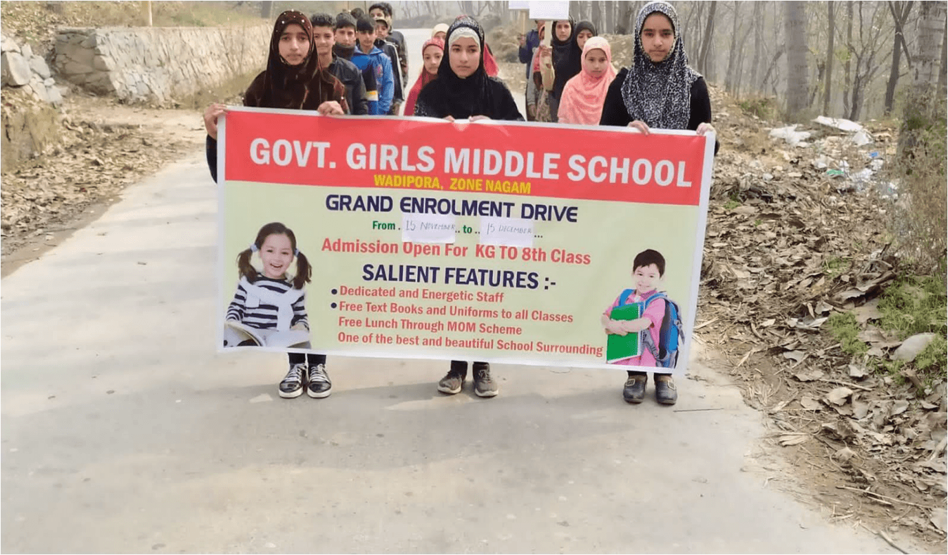 Students walking a village road behind a banner for a Government Girls Middle School enrolment drive