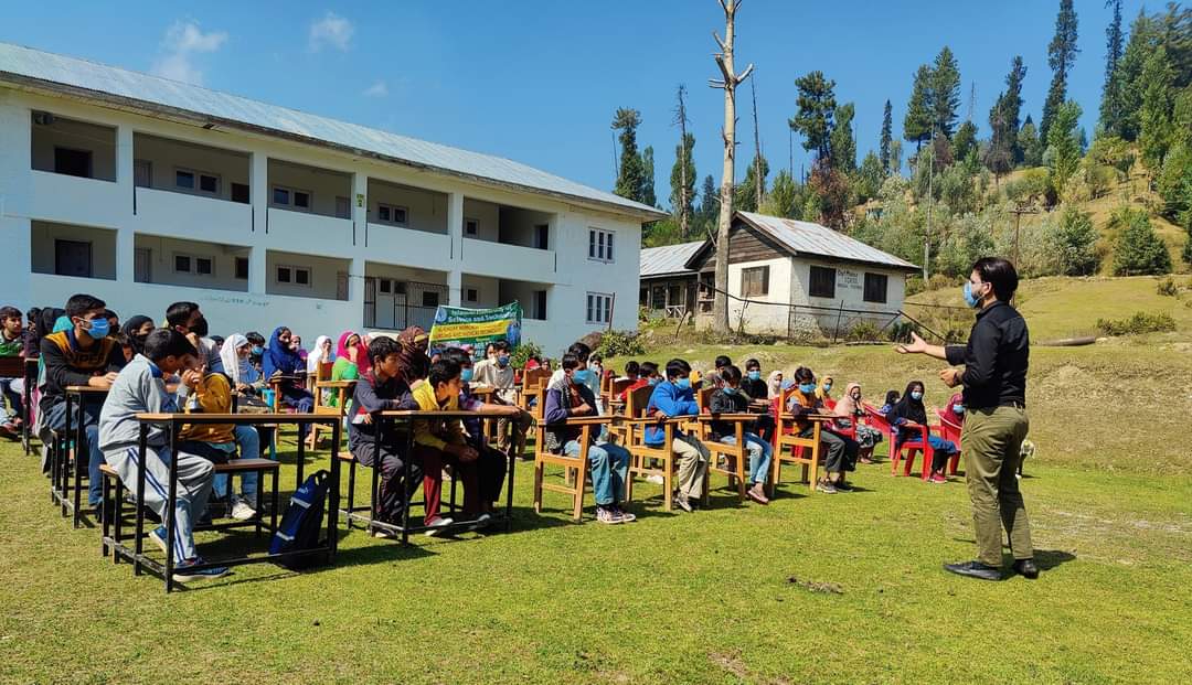 A facilitator addressing students seated at desks on a school lawn in a hillside village